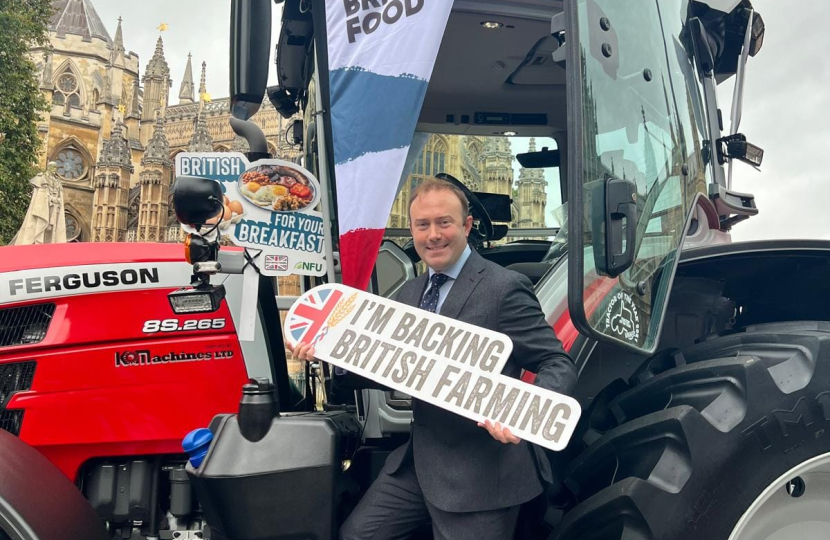 Blake Stephenson MP in a grey suit holding a sign reading 'I'm Backing British Farming' whilst stood on the steps of a red tractor outside Parliament