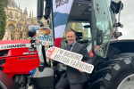 Blake Stephenson MP in a grey suit holding a sign reading 'I'm Backing British Farming' whilst stood on the steps of a red tractor outside Parliament