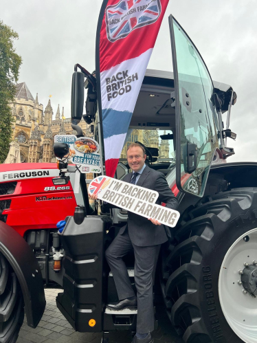 Blake Stephenson MP in a grey suit holding a sign reading 'I'm Backing British Farming' whilst stood on the steps of a red tractor outside Parliament